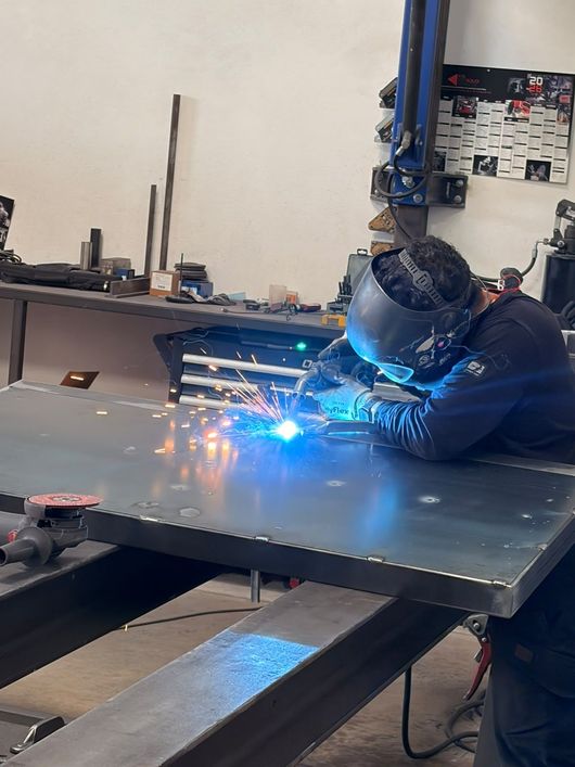 Dans un atelier, un soudeur utilise un chalumeau sur une table métallique, projetant des étincelles d'un bleu vif.