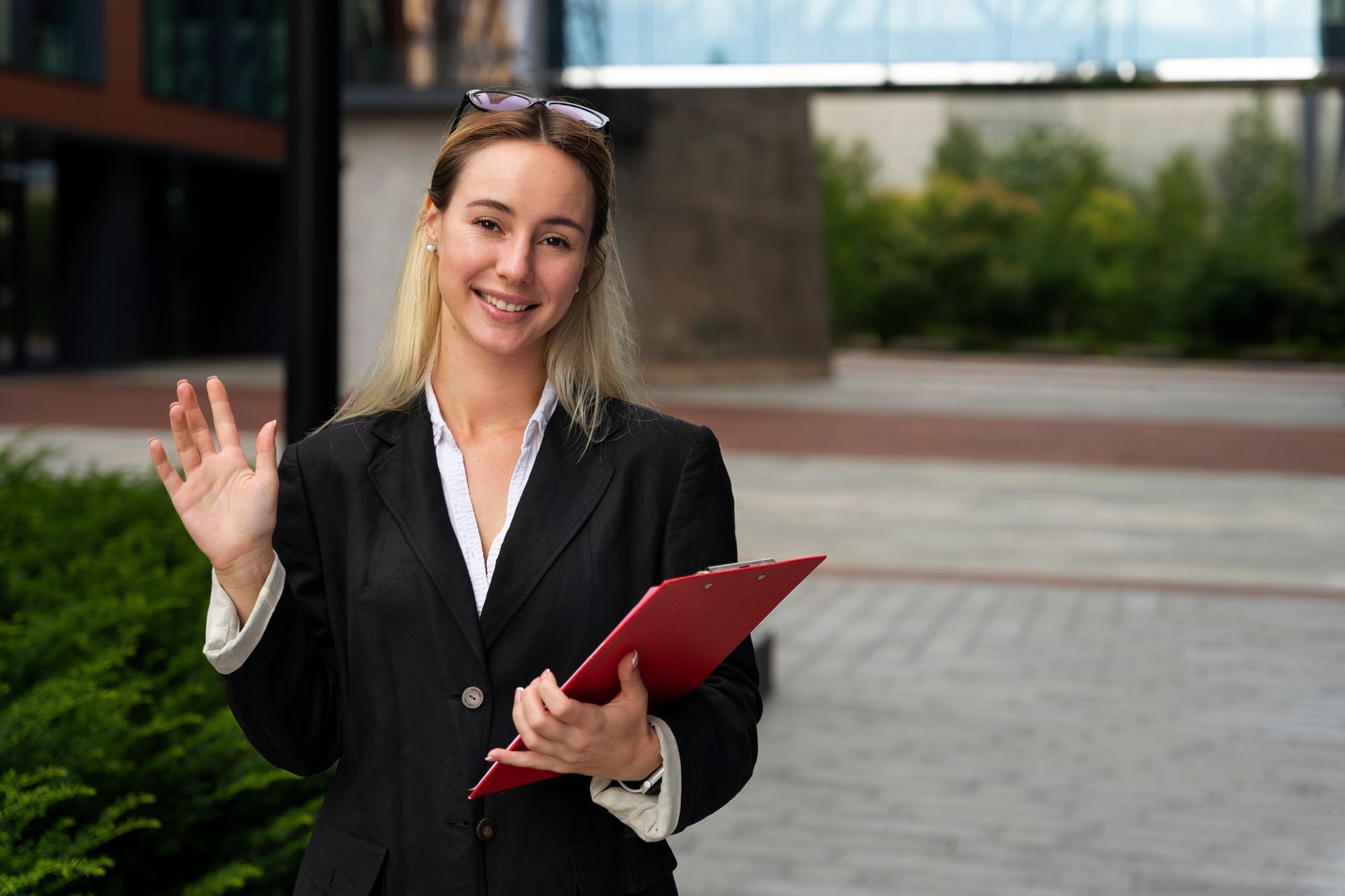 Une femme en blazer noir fait un signe de la main, tenant un bloc-notes rouge, souriante, à l'extérieur près d'un bâtiment.