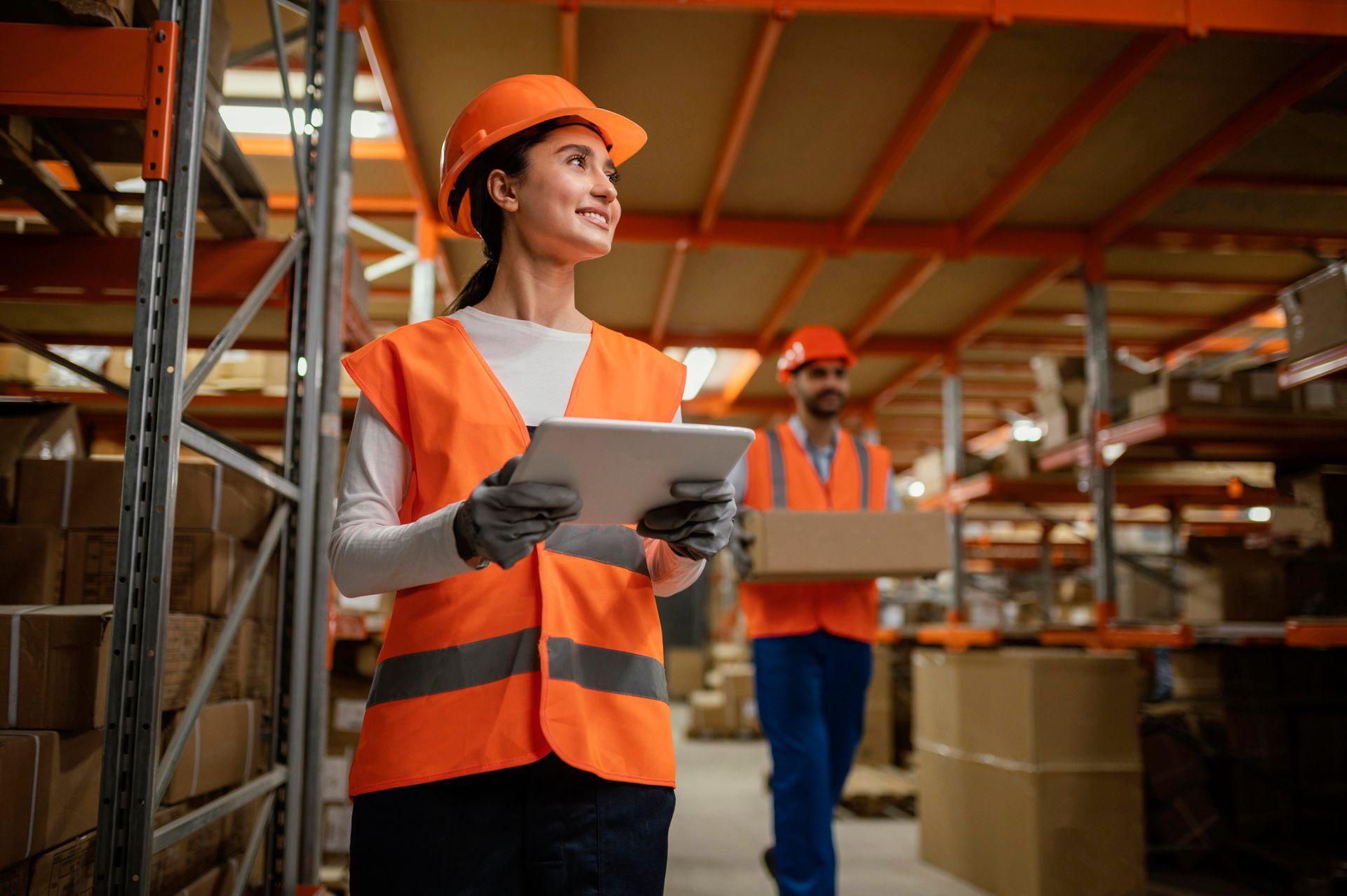 Une femme portant un gilet de sécurité et un casque de chantier utilise une tablette dans un entrepôt.