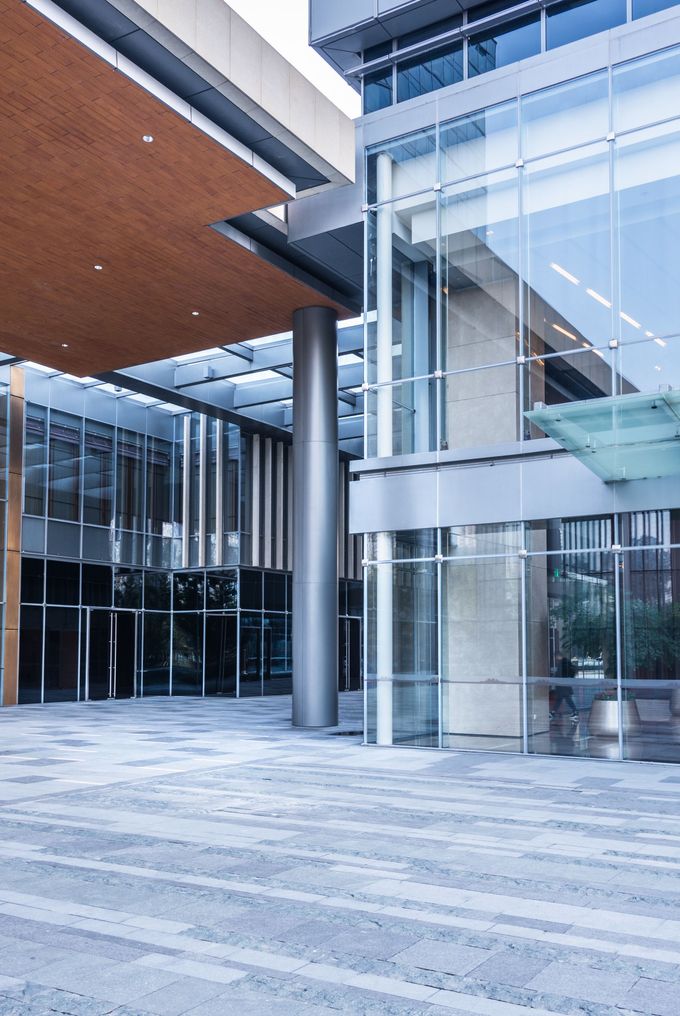 Façade d'un bâtiment moderne avec murs de verre, colonne en acier et sol en pierre à motifs.