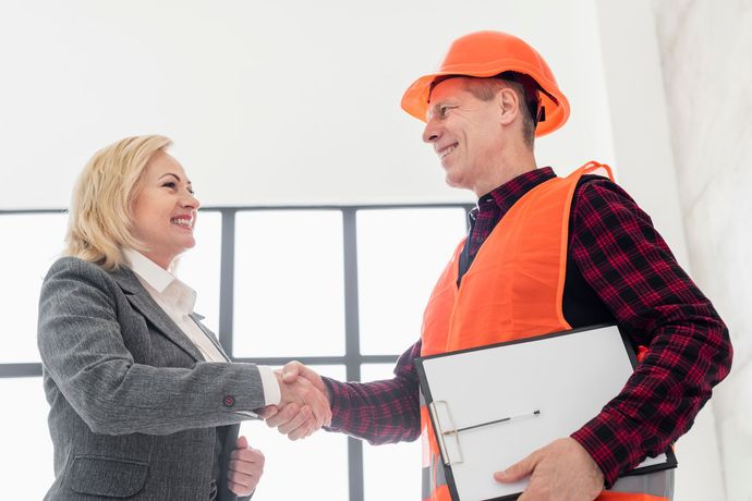 Une femme en tailleur serre la main d'un ouvrier du bâtiment portant un gilet de sécurité et un casque de chantier.