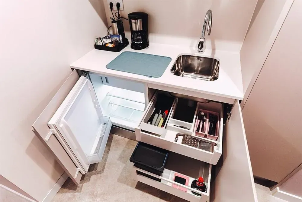 Pequeña cocina con nevera, fregadero, cafetera y cajones llenos de suministros.