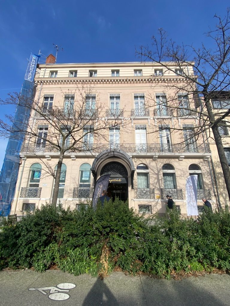 Immeuble beige à plusieurs étages avec entrée cintrée, balcons et arbres dénudés se détachant sur un ciel bleu.