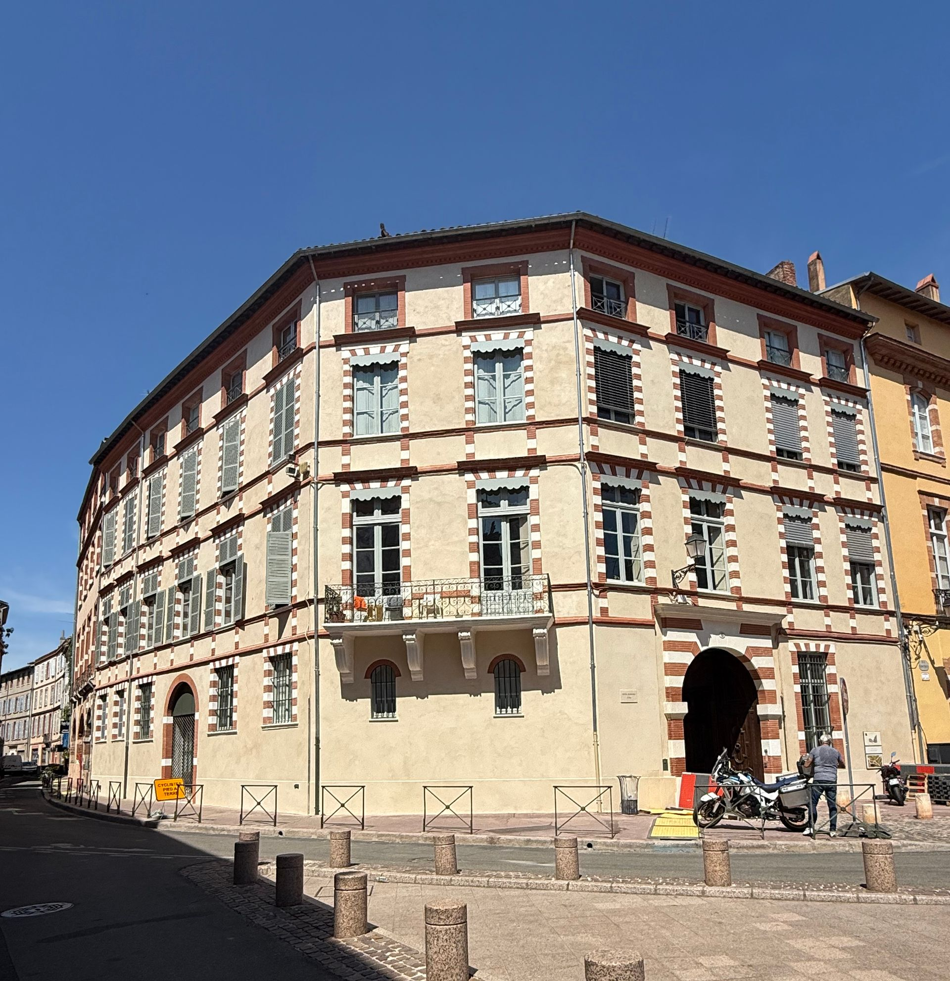 Immeuble de trois étages à la façade beige, aux boiseries rouges et à l'entrée cintrée, situé à un angle de rue, sous un ciel bleu.