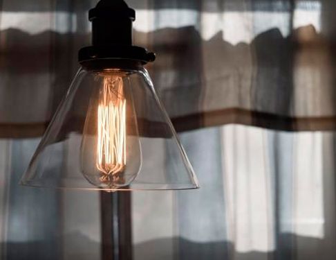 Bombilla Edison encendida bajo una pantalla de cristal frente a unas cortinas transparentes.