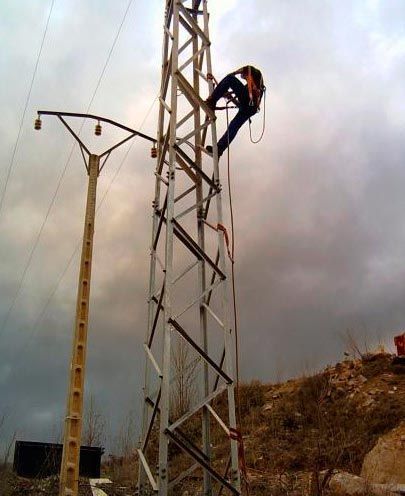 Un trabajador trepa a una alta torre eléctrica de metal junto a postes de servicios públicos bajo un cielo nublado.