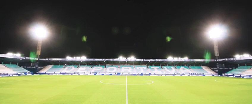 Estadio de fútbol vacío por la noche, con potentes focos iluminando el campo verde y las gradas.