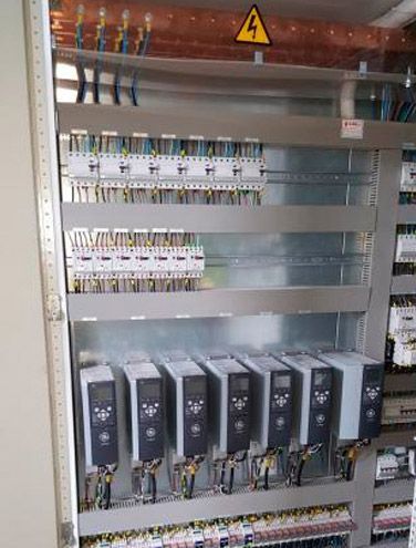 Panel de control eléctrico con filas de disyuntores, cableado y una señal amarilla de advertencia de alto voltaje.