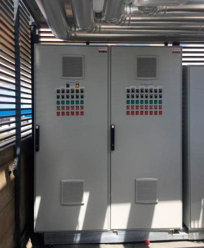 Armarios de control eléctrico grises con etiquetas de advertencia en una sala industrial con tuberías de techo a la vista.