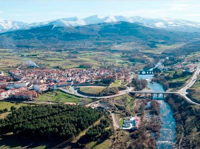 Vista aérea de un pueblo junto a un río con puentes, campos y montañas en la distancia.