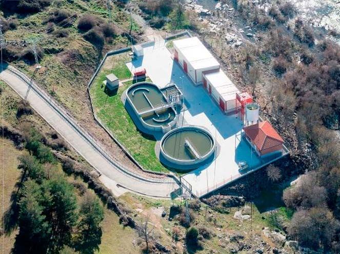 Vista aérea de una planta de tratamiento de agua con tanques redondos, edificios y colinas rocosas circundantes.