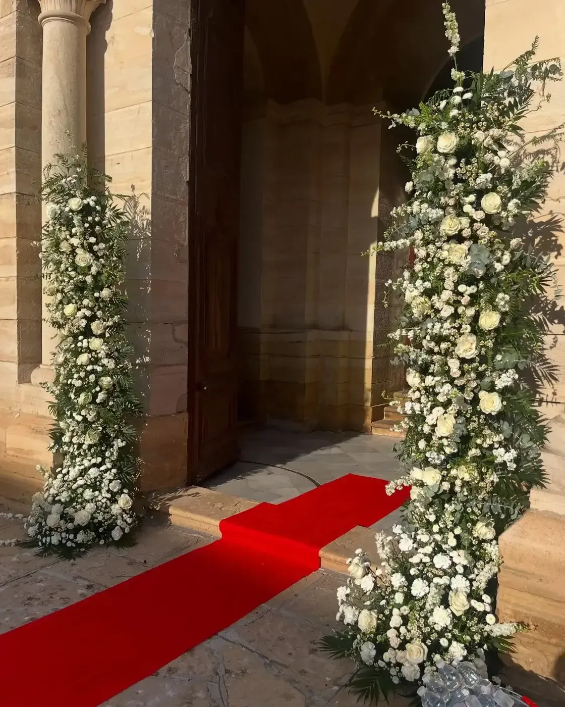 Entrada con alfombra roja a un edificio enmarcado por arreglos florales de flores blancas y vegetación.