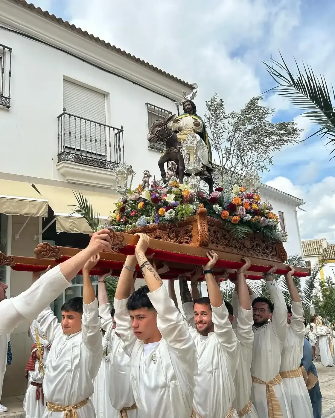Personas portando una plataforma decorada con una estatua, posiblemente en una procesión religiosa. Edificio blanco al fondo.
