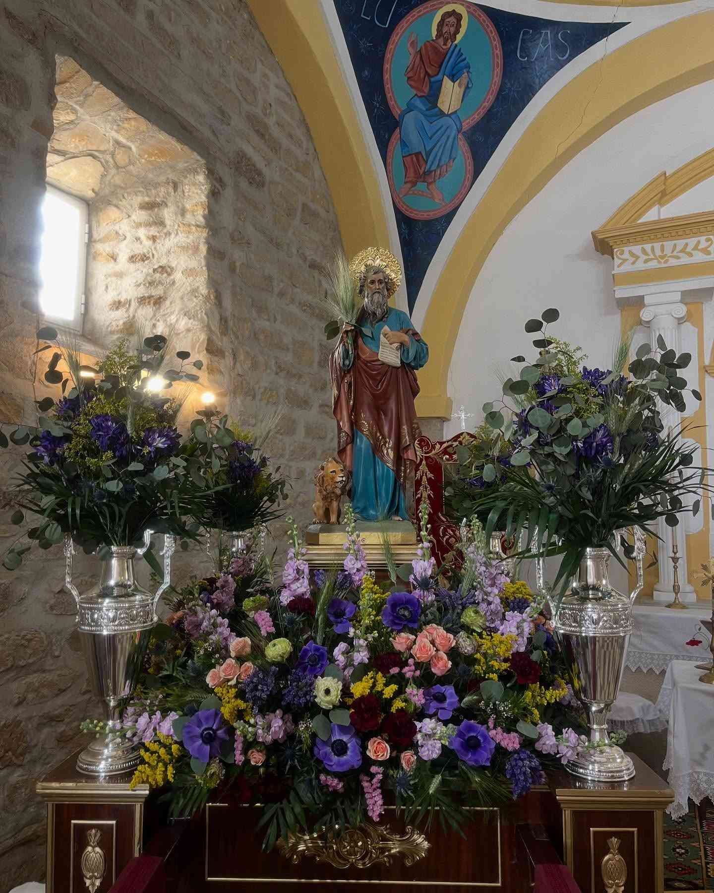 Estatua de un santo rodeado de flores en una iglesia. Se puede ver un mural en la parte superior.