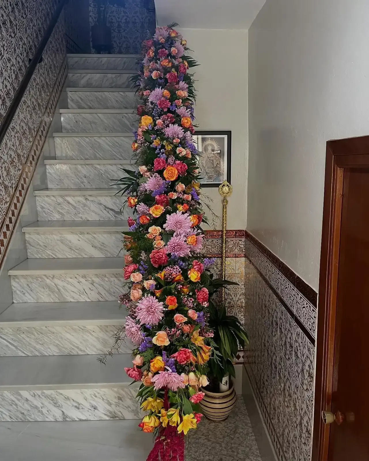 Escalera decorada con flores de colores. Un retrato cuelga de la pared y una planta se encuentra cerca del final.