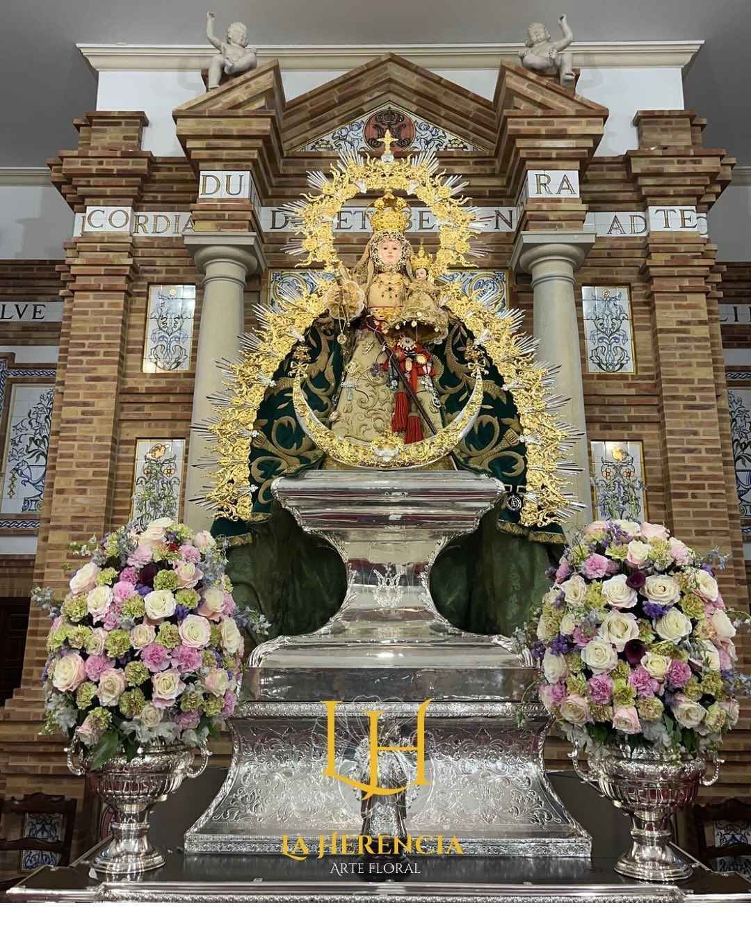Estatua de la Virgen María con el Niño Jesús, plataforma plateada ornamentada, flores. Fondo de ladrillo y columnas.
