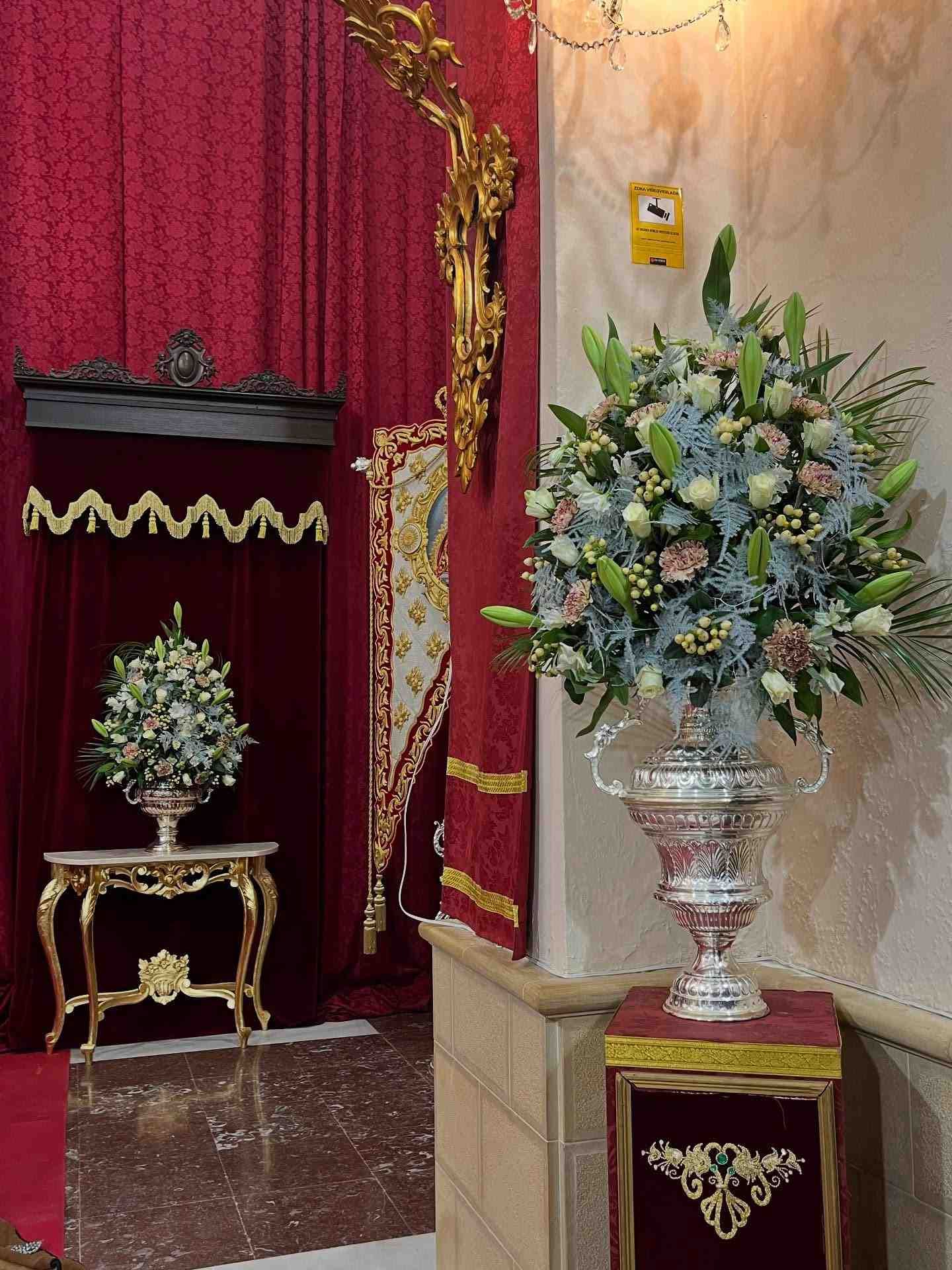 Dos grandes arreglos florales en urnas de plata, uno sobre una mesa dorada. Cortinas rojas y detalles ornamentales.