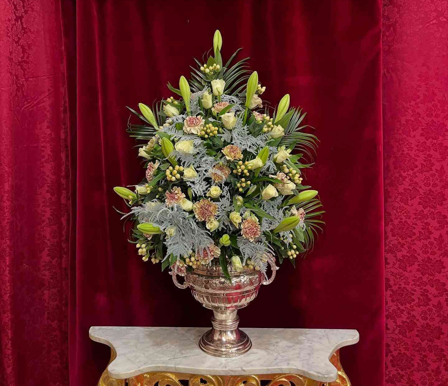 Arreglo floral en jarrón de plata sobre una mesa ornamentada contra una cortina roja.
