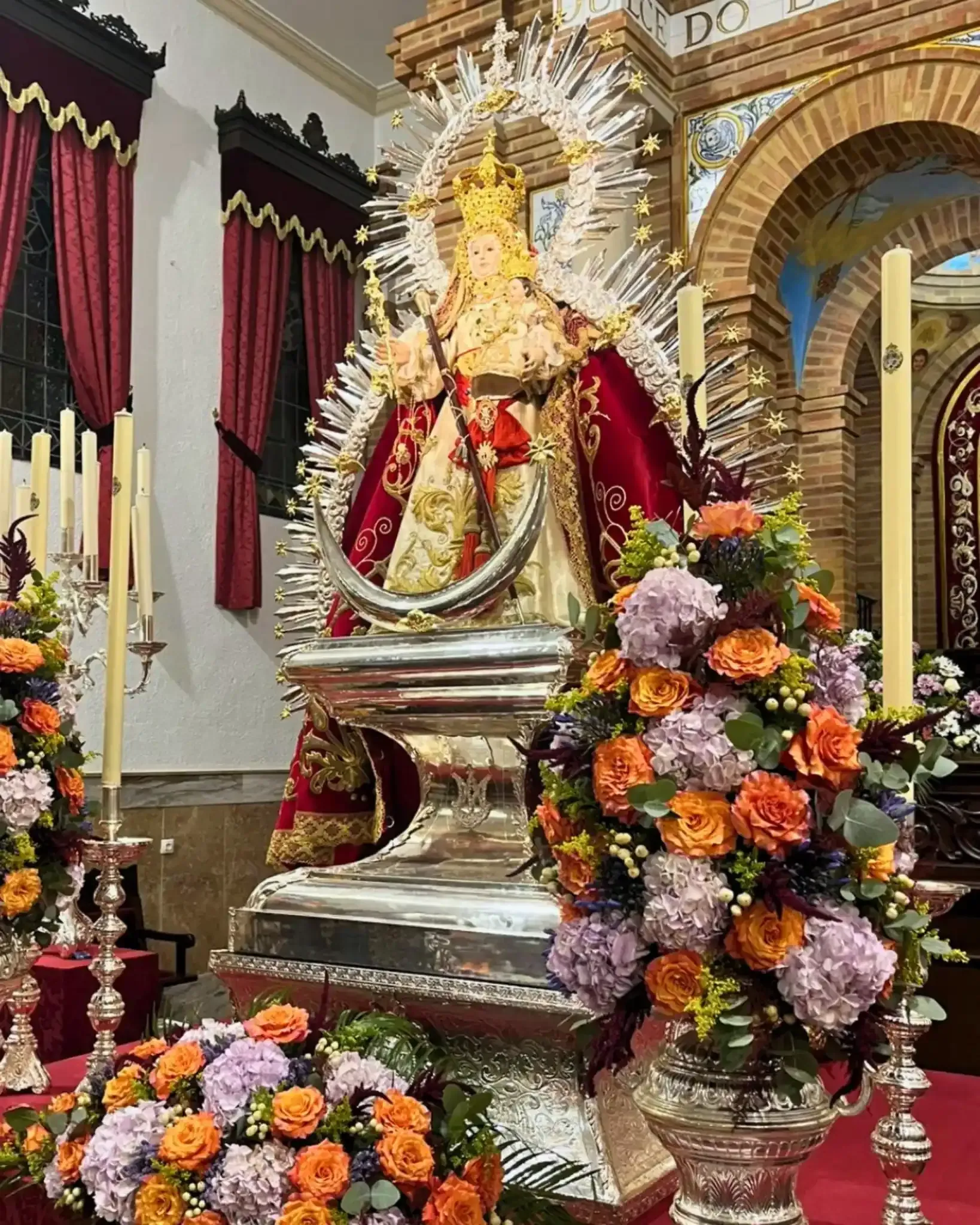 Estatua de la Virgen María en una iglesia, rodeada de flores y velas.