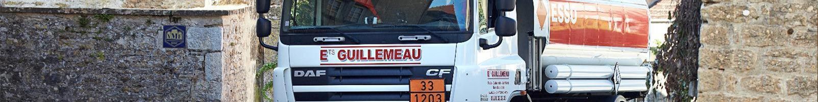 Vue plongeante d'un camion-citerne blanc à réservoir orange, dans une ruelle étroite entre des murs de pierre.