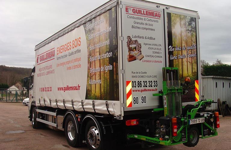 Un camion blanc avec des publicités pour des entreprises liées au bois et une nacelle élévatrice verte à l'arrière.