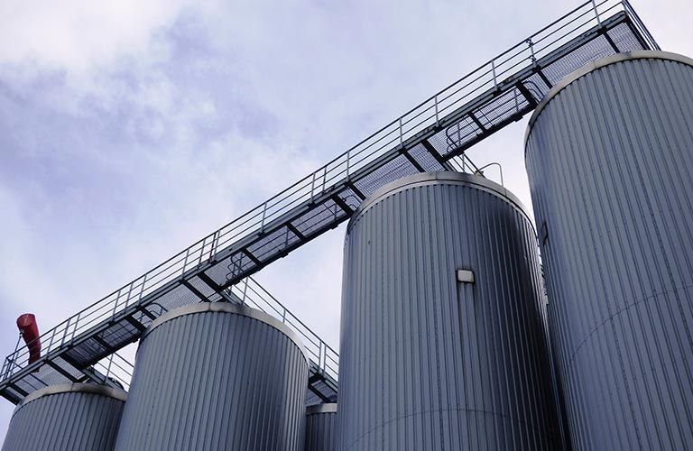 Réservoirs de stockage industriels gris reliés par une passerelle métallique, sur fond de ciel nuageux.