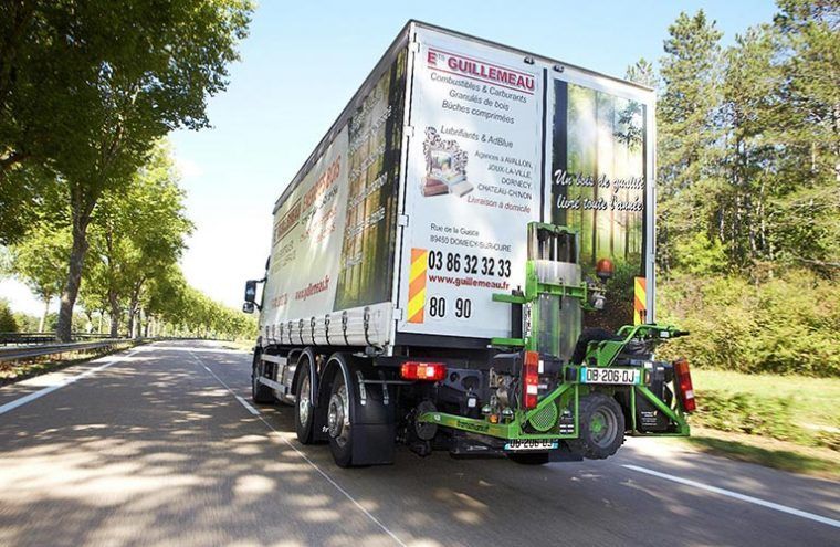 Camion circulant sur la route, tractant une remorque avec une machine attelée. Couleurs vertes et blanches, arbres en arrière-plan.