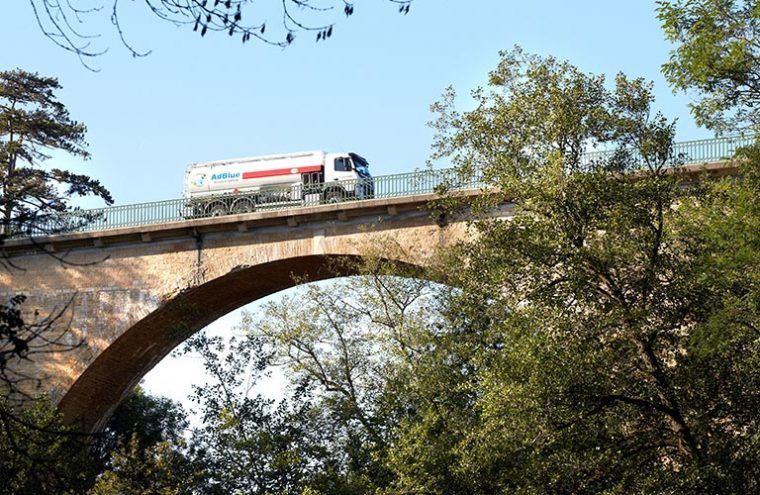 Camion-citerne traversant un pont en arc de pierre, arbres au premier plan, ciel dégagé.