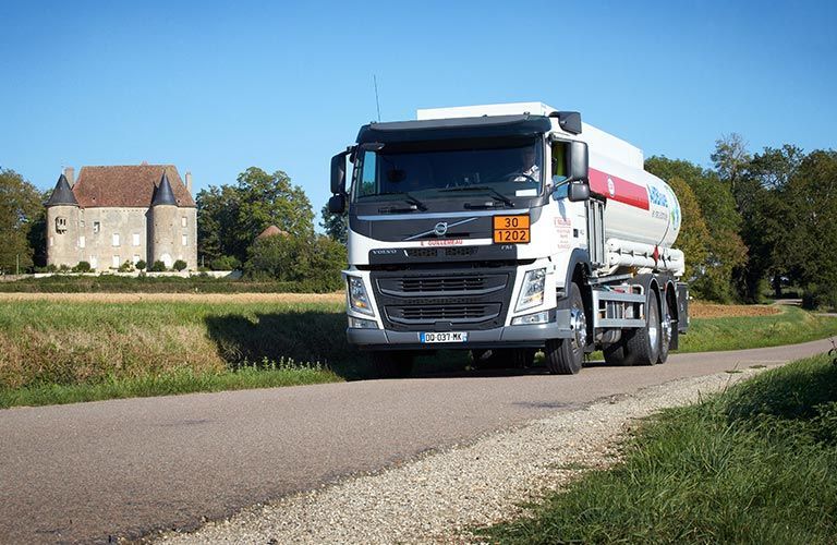 Camion-citerne sur une route, avec un bâtiment et des champs en arrière-plan par une journée ensoleillée.