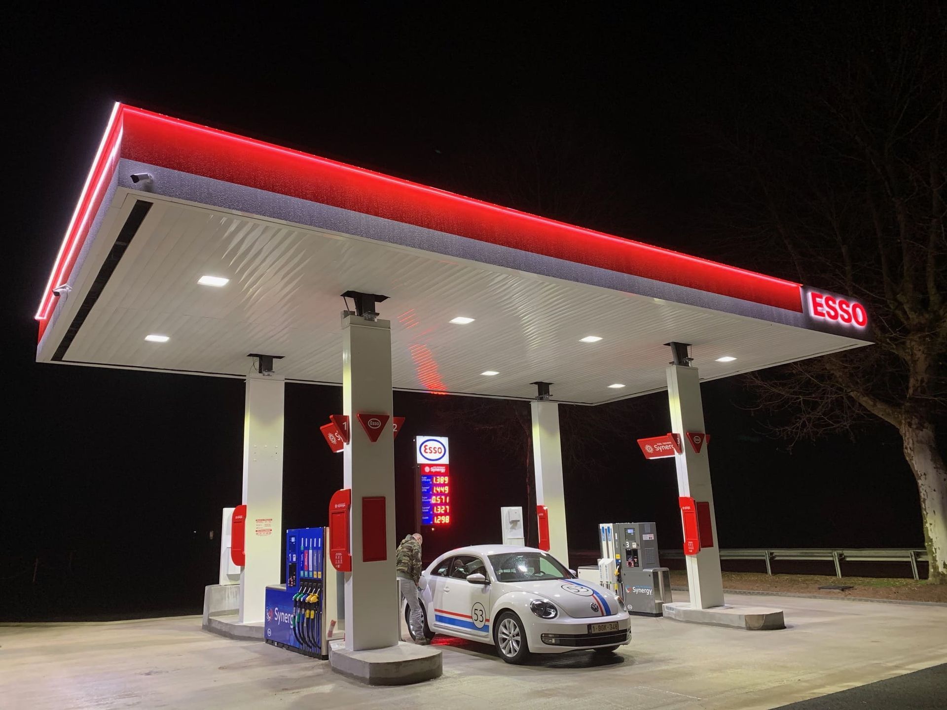 Station-service Esso la nuit, avec une voiture blanche en train de faire le plein. Auvent rouge et blanc.