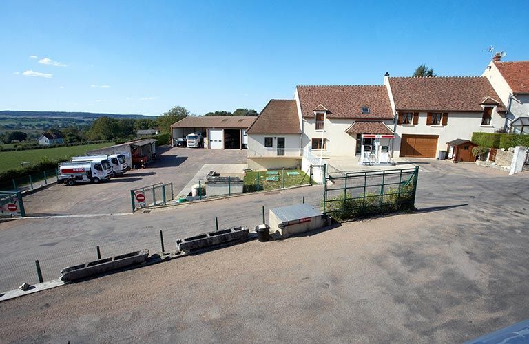 Vue diurne ensoleillée d'un complexe de bâtiments ruraux avec un parking, des véhicules et une clôture basse.