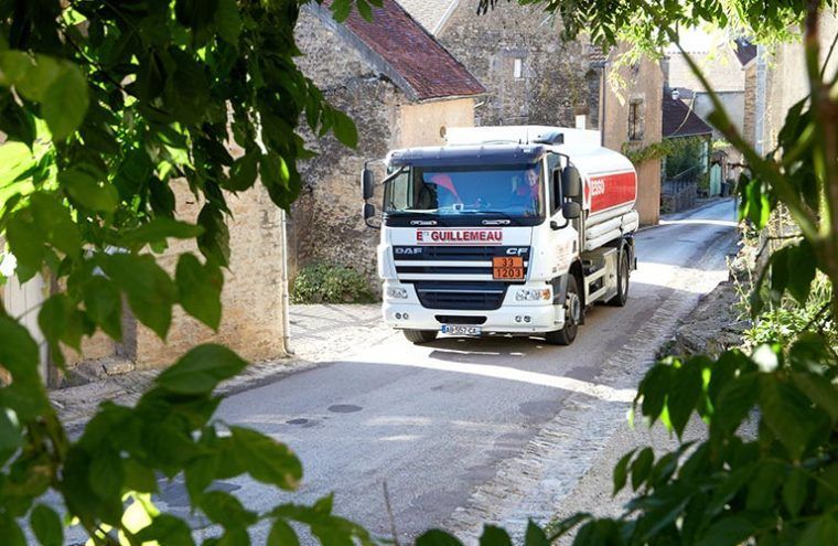 Un camion-citerne circule dans une rue pavée étroite d'un village, encadrée par un feuillage vert.