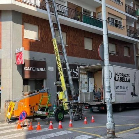 Un camión de mudanzas y un ascensor junto a un edificio, conos en la calle. Letrero de 