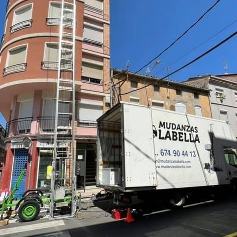 Camión de mudanzas con elevador, estacionado en la calle frente a un edificio. Logotipo de 