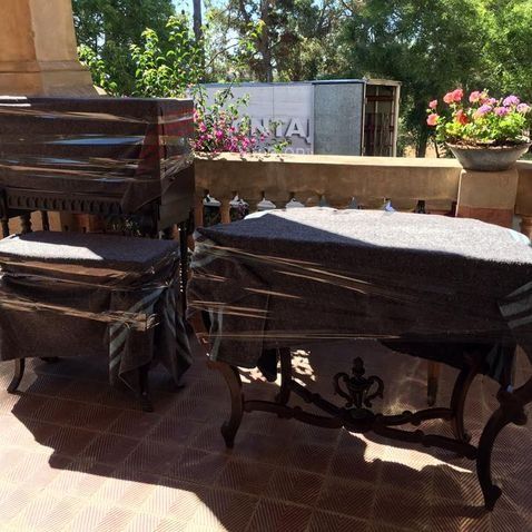 Dos sillas de madera antiguas y una mesa están envueltas en plástico en un patio exterior de baldosas.