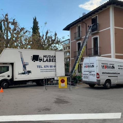 Camiones de mudanzas con un elevador que se extiende hasta un edificio de apartamentos; señal amarilla de “no estacionar”.