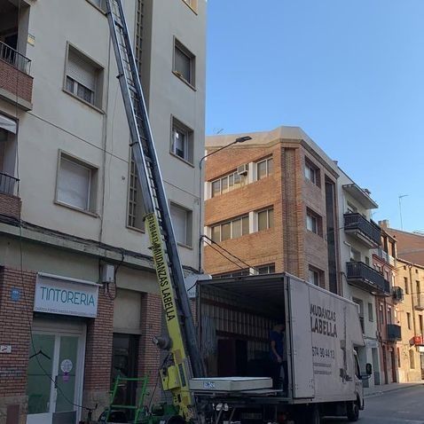Un camión de mudanzas con elevador se estanca frente a un edificio de apartamentos. Un trabajador carga un colchón.