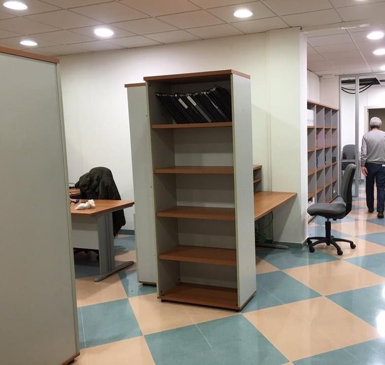 Interior de oficina con estantería, escritorios y suelo de cuadros. Una persona camina al fondo.