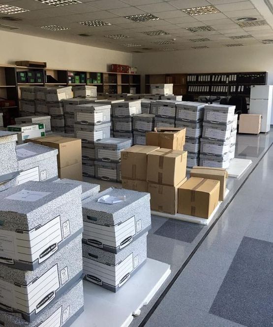 Cajas apiladas en una habitación grande. Las cajas son grises, marrones y canela. Se ve un refrigerador.