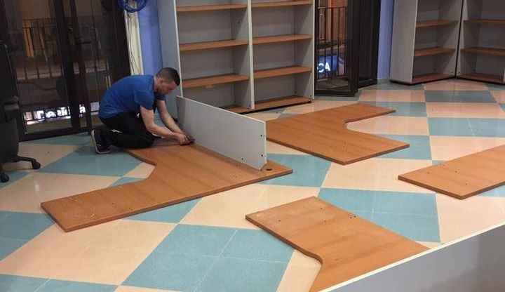 Una persona ensambla muebles de oficina sobre un piso cuadriculado, con estanterías en el fondo.