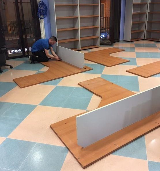 Una persona ensambla un componente de escritorio sobre un piso cuadriculado, con estanterías en el fondo.