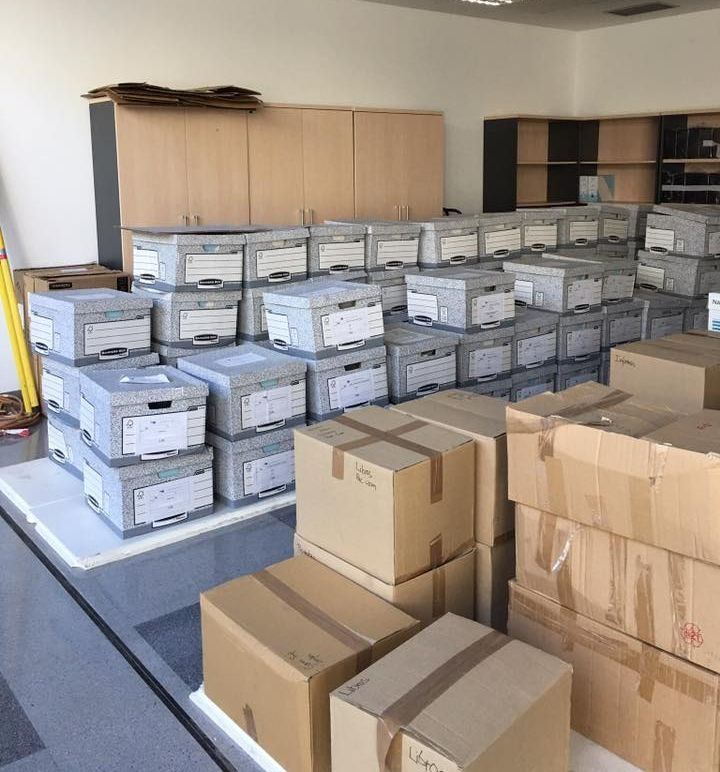 Cajas de archivos apiladas en una sala de almacenamiento con armarios al fondo.