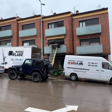 Camiones de mudanzas y un Jeep estacionados frente a un edificio; muebles siendo trasladados a un balcón.