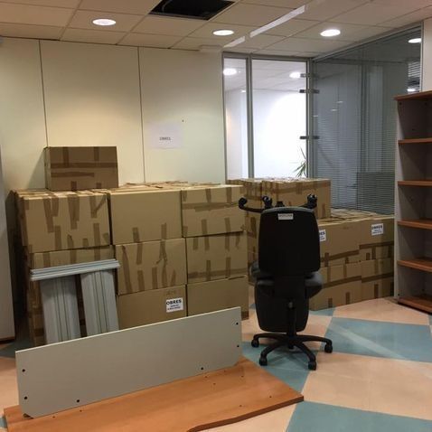Espacio de oficina en desorden; cajas apiladas, sillas, muebles desmontados y estantes vacíos.