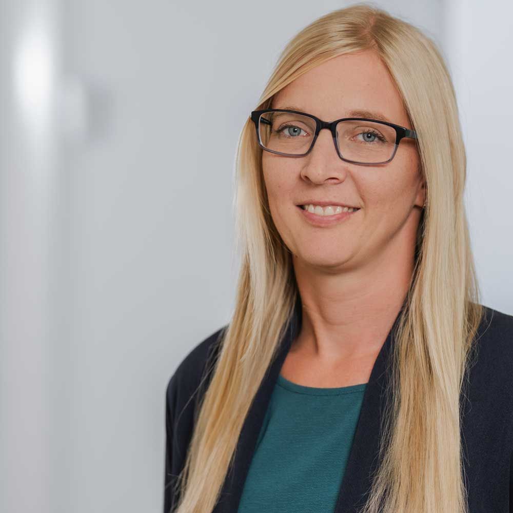 Eine Frau mit langen blonden Haaren, Brille und blauer Jacke lächelt in einem hellen Flur.
