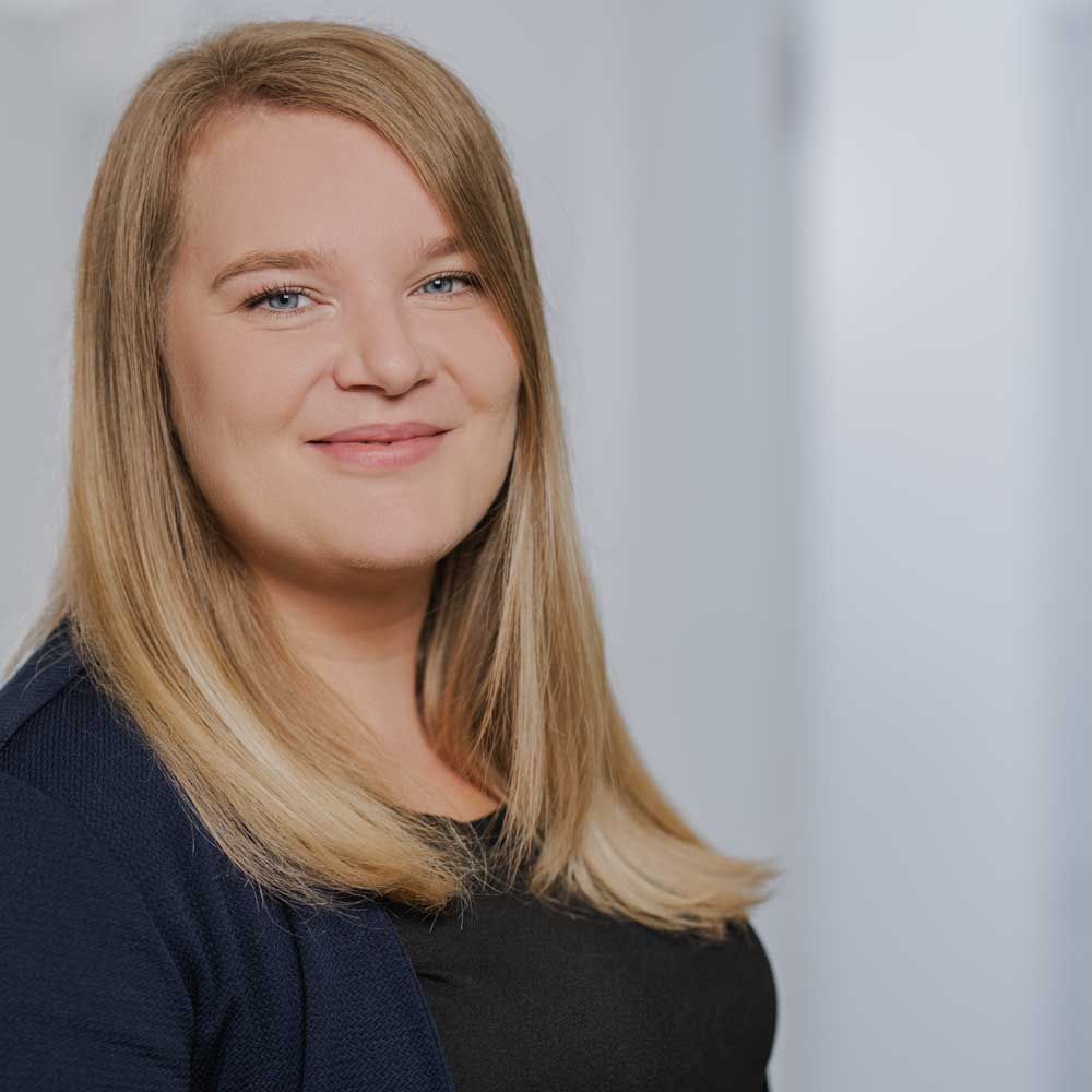 Eine lächelnde Frau mit blonden Haaren, die eine dunkelblaue Jacke und ein schwarzes Oberteil trägt, in einem gut beleuchteten Büro.