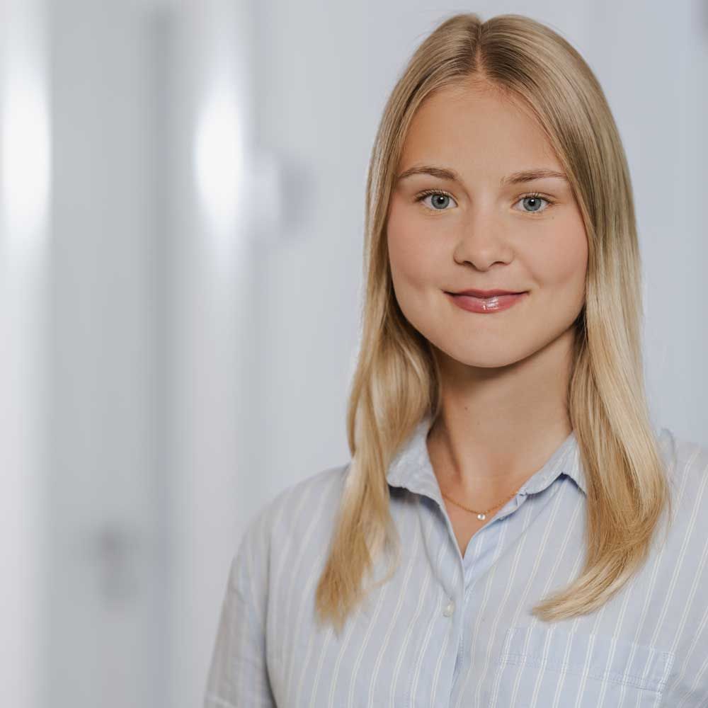 Blonde Frau lächelt in einem hellblau gestreiften Hemd in einem hellen Büro.