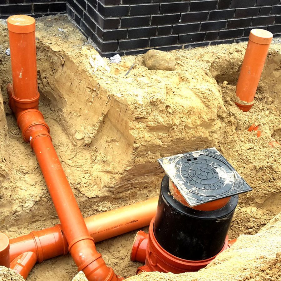 Orangefarbene PVC-Rohre und ein schwarzer Abwasseranschluss wurden in einem sandigen Graben in der Nähe einer Ziegelmauer installiert.