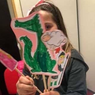 Una niña sostiene un dibujo de un cocodrilo en un palo frente a su cara.