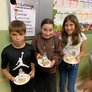 Un niño y dos niñas sostienen platos de comida delante de un cartel de bienvenida.